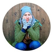 Frau trägt eine graue Mütze, Handschuhe und eine grüne Jacke, sitzt vor einem Holzzaun, wirkt nachdenklich und entspannt.