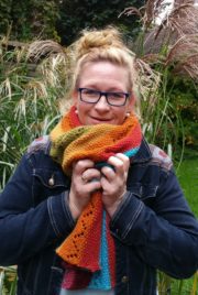 Frau mit buntem Strickschal, Brille und Jeansjacke, umgeben von Gräsern und Pflanzen, zeigt kreative Handarbeit und Naturverbundenheit.