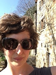 Frau mit kurzen braunen Haaren und Sonnenbrille, im Hintergrund eine alte Steinmauer und blauer Himmel, bei sonnigem Wetter, Fokus auf das Gesicht.