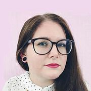 Frau mit Brille, dunklem Haar und roten Lippen, trägt eine weiße Bluse mit schwarzen Punkten, vor einem helllila Hintergrund, repräsentiert kreative Handwerkskunst und Kultur.