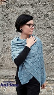 Frau trägt einen handgefertigten, blauen Strickschal, der kunstvoll gefaltet ist, mit schwarzer Kleidung und Brille, vor einer Steinmauer, ideal für Handmade- und Strickkunst-SEO.