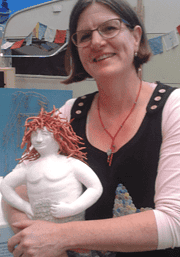 Frau mit Brille und Kurzhaarfrisur zeigt eine handgefertigte Skulptur eines nackten Jungen mit roten Haaren, umgeben von bunten Stoffflaggen im Hintergrund.