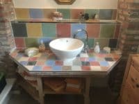 Badezimmer mit handgefertigtem Fliesendesign, bunte quadratische Fliesen in verschiedenen Farben, Waschbecken und Badezimmeraccessoires, rustikaler Charme, kreative Raumgestaltung.