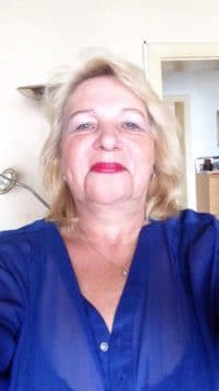 Porträt einer Frau mit blonden Haaren, rotem Lippenstift und blauer Bluse, die für Handmade Kultur steht, in einem häuslichen Umfeld.