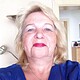Porträt einer Frau mit blonden Haaren, rotem Lippenstift und blauer Bluse, die für Handmade Kultur steht, in einem häuslichen Umfeld.