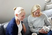 Zwei lachende Frauen sitzen auf einem Sofa, trinken Kaffee und genießen einen entspannten Moment zusammen.