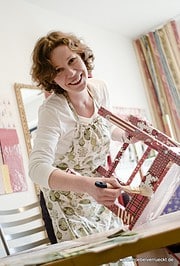 Junge Frau beim kreativen Malen mit Pinsel und Farben, fröhlich und engagiert, in einem hellen Raum mit bunten Vorhängen, fördert kreative Handwerkskunst und DIY-Projekte.
