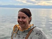 Frau mit braunen Haaren, grauem Hoodie mit Hirschmotiven, lächelnd, am Wasser mit Bergen im Hintergrund, kreative Handarbeit, nachhaltige Mode, deutsches Handmade-Design.