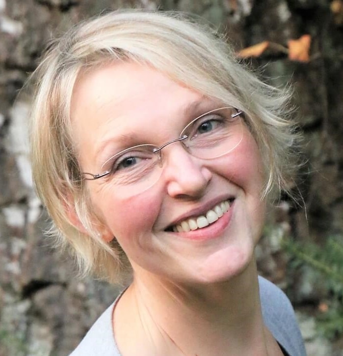 Lächelnde Frau mit Brille und kurzen blondem Haar, umgeben von Natur, symbolisiert kreative Handwerkskunst und nachhaltiges Handarbeiten bei Handmade Kultur.