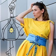 Frau in gelbem Kleid mit blauer Gürtelschnalle, kreative Frisur, neben einer handgezeichneten Cartoon-Figur, symbolisiert Handgemacht und Kreativität.