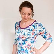 Frau trägt ein buntes Blumenmuster-Shirt mit 3/4-Ärmeln, das handgefertigt ist, und posiert vor neutralem Hintergrund für ein kreatives Modefoto.