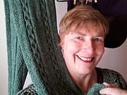 Lächelnde Frau mit kurzen Haaren zeigt handgefertigten, grünen gestrickten Schal, der auf einem Kleiderbügel hängt, bei Handmade Kultur.