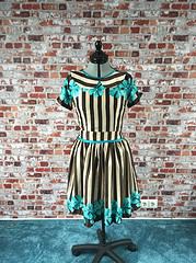 Handgefertigtes Kleid mit schwarzen und weißen Streifen, blauen Blumenakzenten, kurzer Ärmel, auf einem Modenschau-Stand vor Ziegelwand präsentiert.