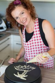 Frau beim kreativen Backen in der Küche, trägt eine rote und weiße Schürze, lächelt freundlich, bereitet Kekse oder Dekorationen vor, zeigt handwerkliches Geschick.