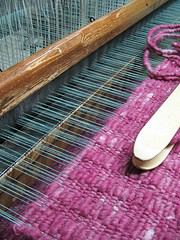 Detailaufnahme eines handgefertigten Teppichs auf einem Webstuhl, zeigt die Webtechnik und die Textur des Teppichs in pinker Farbe.