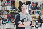 Freundliche Frau in Handwerksladen mit buntem Sortiment an Wolle, Stoffen und Bastelmaterialien, die handgemachte Produkte präsentiert.