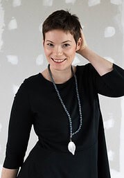 Frau mit kurzen Haaren trägt schwarze Kleidung und eine lange Kette mit Anhänger, lächelnd, vor einem weißen Wand mit Pinselstrichen, kreative Atmosphäre.