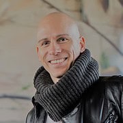 Mensch mit schwarzem Lederjacke und grauem Schal, lächelnd, in einem kreativen Handwerksumfeld, das handgemachte Produkte und Kunst zeigt.