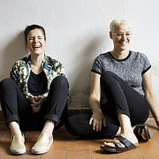 Zwei Frauen sitzen entspannt auf dem Boden, lachen und genießen kreative Handwerkskunst bei Handmade Kultur, einer Plattform für handgemachte Produkte und kreative Gemeinschaften.