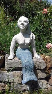 Kunstvolle Handmade Skulptur einer sitzenden Frau aus Stein, umgeben von bunten Blumen und grüner Natur, ideal für Garten- und Kunstliebhaber.