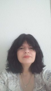 Porträt einer Frau mit dunklem, lockigem Haar und neutralem Gesichtsausdruck, vor weißer Wand, in casual Kleidung, natürliche Beleuchtung, Fokus auf Gesicht und Haare.