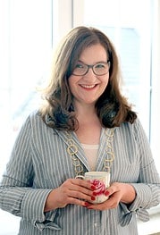 Frau mit Brille und Lächeln hält eine Tasse mit Blumenmuster, trägt eine gestreifte Bluse und eine goldene Kette, im Hintergrund ein Fenster mit natürlichem Licht.