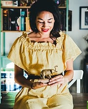 Frau hält eine kleine Elefantenfigur aus Metall, umgeben von einem kreativen, handgemachten Ambiente mit Büchern und Dekorationen im Hintergrund.