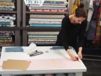 Frau beim Zuschneiden von Stoffen in einem Stoffladen, umgeben von bunten Stoffrollen und Nähzubehör, für handgemachte Textilkunst und kreative Nähprojekte.