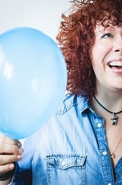 Lächelnde Frau mit lockigem Haar hält einen blauen Ballon, symbolisiert Freude und Feierlichkeit, passend für kreative und handgemachte Veranstaltungen.