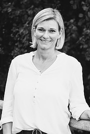 Frau mit kurzen blondem Haar, lächelnd, in weißem Hemd, umgeben von Natur, symbolisiert handgemachte Kreativität und nachhaltiges Handwerk.
