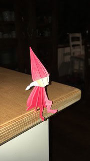 Kleine handgefertigte Weihnachtsfigur aus Papier mit rotem Kleid und weißem Schal, sitzend auf einer Holzoberfläche, ideal für festliche Dekorationen.