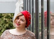 Junge Frau mit rotem Hut und weißem Haar, lächelnd, vor einer Metallzaun, symbolisiert kreative Handwerkskunst und nachhaltiges Design bei Handmade Kultur.