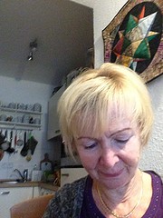 Porträt einer älteren Frau mit blondem Haar, die in einem gemütlichen Raum sitzt, umgeben von handgefertigten Kunstwerken und kreativen Dekorationen.