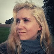 Junge Frau mit blonden Haaren und dunklem Schal blickt nach rechts, im Hintergrund Bäume und offenes Feld, natürliche Lichtverhältnisse.