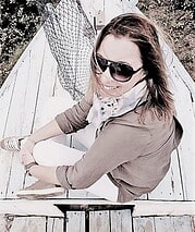 Frau mit Sonnenbrille, casual Kleidung und weißer Hose sitzt auf einer Holzbrücke, umgeben von Natur, bei Handmade Kultur, Fokus auf kreative Handwerkskunst und Nachhaltigkeit.
