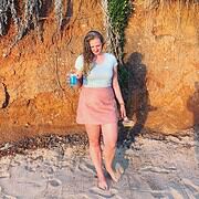 Junge Frau mit langem Haar trägt rosa Rock und weißes T-Shirt, hält Kaffee und spaziert barfuß am Strand entlang, im Hintergrund Felsen und Sand.