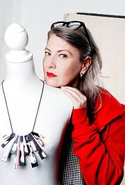 Frau mit roten Lippen und grauem Haar posiert neben einer Schmuckpuppe, trägt eine rote Jacke und zeigt ein modernes, handgemachtes Schmuckstück aus recyceltem Material.