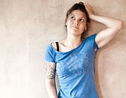 Junge Frau mit Tattoo und blauer T-Shirt wirkt nachdenklich bei HANDMADE Kultur, kreative Atmosphäre, handgemachte Kunst, Inspiration, Kunsthandwerk, DIY, kreative Gemeinschaft.