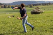 Frau spielt mit Hunden im offenen Feld, umgeben von Wiesen und Bäumen, bei HANDMADE Kultur, Natur, Tierliebe, Outdoor-Aktivitäten, Gemeinschaft, Freizeit, ländliche Umgebung.