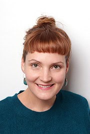 Lächelnde Frau mit roten Haaren, Zopf und blauen Ohrringen, trägt einen blauen Pullover, vermittelt Kreativität und Handwerkskunst.