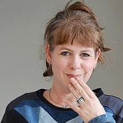Frau mit kurzen braunen Haaren, die nachdenklich lächelt und die Hand vor den Mund hält, trägt eine dunkle Jacke mit blauen Akzenten, kreative Handwerkskunst im Fokus.