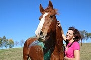 Fröhliche Frau in pinkem T-Shirt streichelt ein braunes Pferd auf einer Wiese, umgeben von Bäumen und blauem Himmel, bei Handmade Kultur.