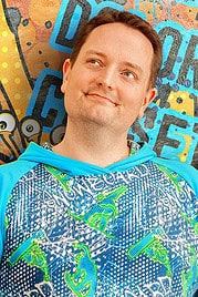 Mann in bunter, handgemachter Kleidung vor bunter Wand mit Graffiti, symbolisiert kreative Handwerkskunst und DIY-Projekte bei Handmade Kultur.