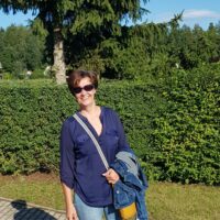 Frau mit Sonnenbrille und blauer Bluse im Garten, umgeben von grünen Büschen und Bäumen, bei sonnigem Wetter, für Handmade Kultur SEO optimiert.
