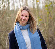 Frau mit blauer Schal und grauem Pullover in Waldumgebung, strahlendes Lächeln, natürliche Atmosphäre, Fokus auf authentische Handwerkskunst und Naturverbundenheit.