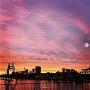 Blick auf die Skyline bei Sonnenuntergang mit farbigem Himmel und Silhouetten von Menschen am Wasser.