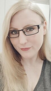 Nahaufnahme einer Frau mit blonden Haaren und schwarzen Brillengestellen, freundliches Gesicht, natürliche Beleuchtung, Fokus auf Gesicht und Augen, professionell und einladend.