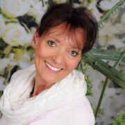 Lächelnde Frau mit kurzen braunen Haaren, trägt weiße Kleidung, umgeben von grünen Pflanzen und handgemachten Dekorationen, zeigt kreative Handwerkskunst bei Handmade Kultur.