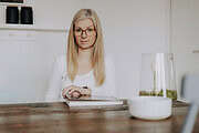 Frau mit Brille und blondem Haar arbeitet an einem Tisch, umgeben von handgemachten Kunstwerken und kreativen Materialien, die handgefertigte Kultur repräsentieren.