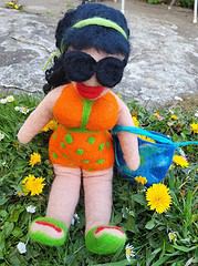 Handgefertigte Stoffpuppe mit dunklen Haaren, Sonnenbrille, orange-grünes Sommerkleid, grüne Schuhe, blauer Strandtasche, im Garten mit Blumen.